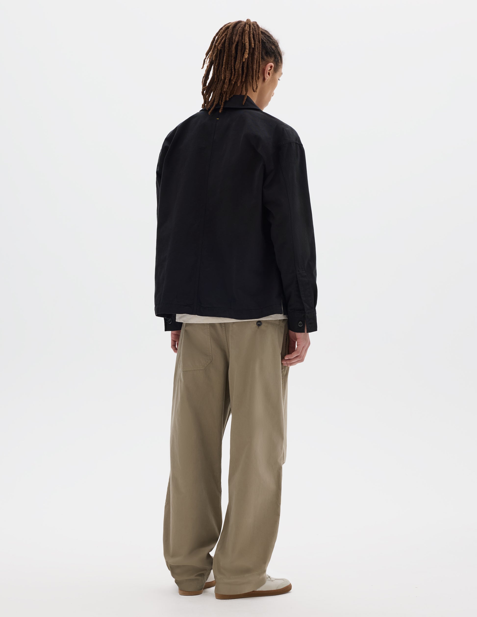 Full body image, back view of model wearing black cotton overshirt, with button closure shirt cuffs and centre back seam. Styled with off white t-shirt, beige trousers and off white trainers with gum sole.