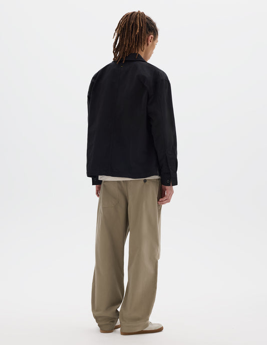 Full body image, back view of model wearing black cotton overshirt, with button closure shirt cuffs and centre back seam. Styled with off white t-shirt, beige trousers and off white trainers with gum sole.