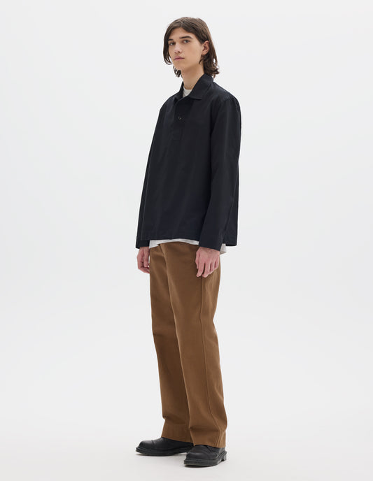 Full body image, front view of model wearing black denim shirt, with wide front placket, two button closure, split placket, and straight side split hem. Styled over white t shirt, with brown trousers and brown leather shoes.
