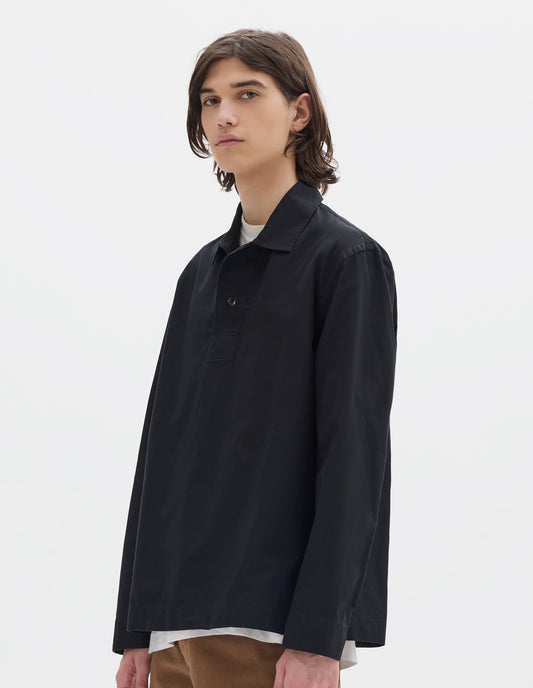 Cropped body image, front view of model wearing black denim shirt, with wide front placket, two button closure, split placket, and straight side split hem. Styled over white t shirt, with brown trousers.