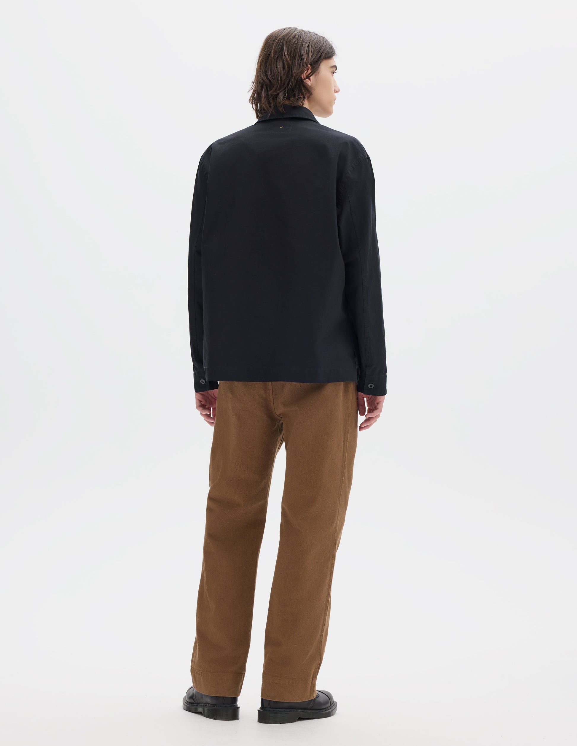 Full body image, back view of model wearing black denim shirt, with button closure cuffs, split placket, and straight side split hem. Styled with brown trousers and brown leather shoes.