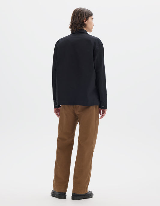 Full body image, back view of model wearing black denim shirt, with button closure cuffs, split placket, and straight side split hem. Styled with brown trousers and brown leather shoes.