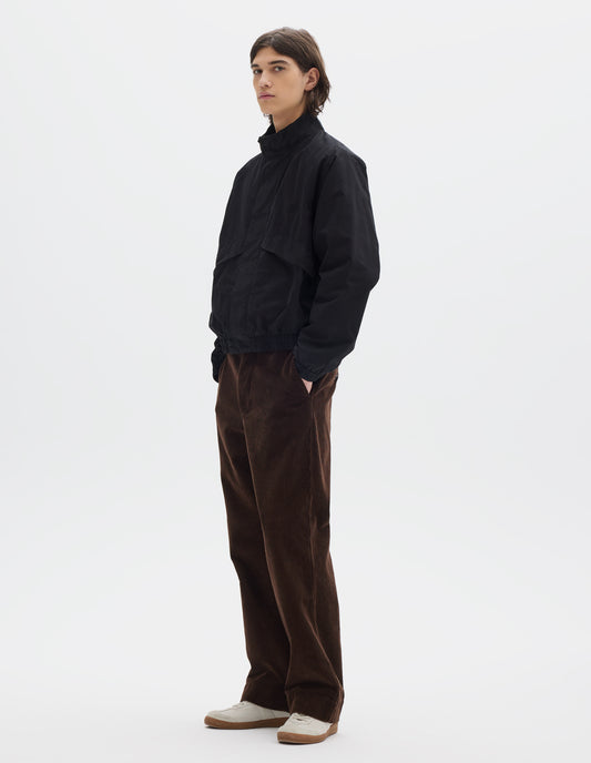 Fully body image, side view of model wearing black cotton poplin track jacket with stand collar, raglan sleeves, concealed zip closure  and press stud fastening and elasticated hem and cuffs. Large slanted front pockets. Styled with brown trousers and white shoes.