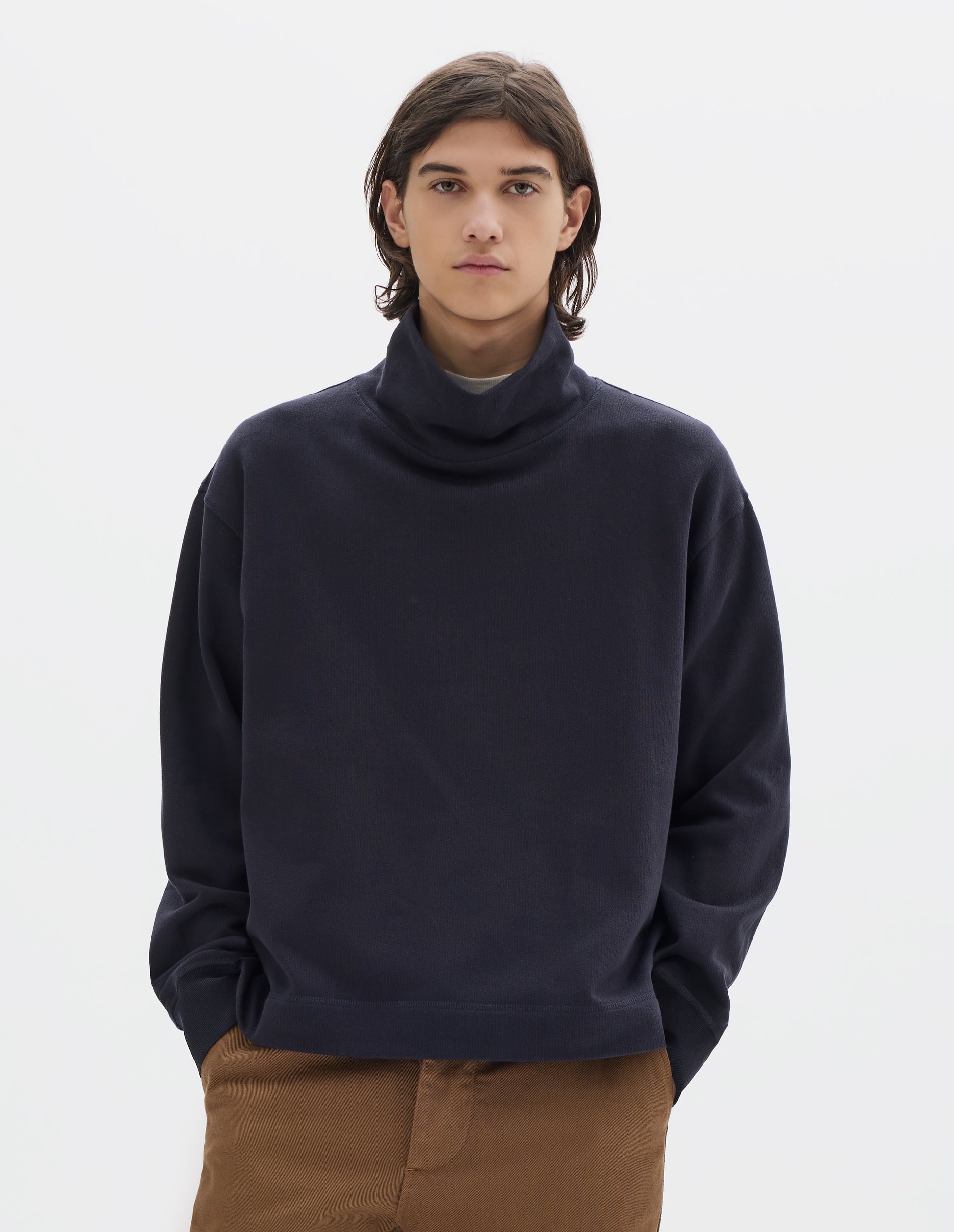 Cropped body image, front view of model wearing a dark navy loose funnel neck sweatshirt with single rib on cuffs and flatlock stitching on seams. Styled with brown cotton trousers.