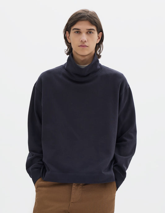 Cropped body image, front view of model wearing a dark navy loose funnel neck sweatshirt with single rib on cuffs and flatlock stitching on seams. Styled with brown cotton trousers.