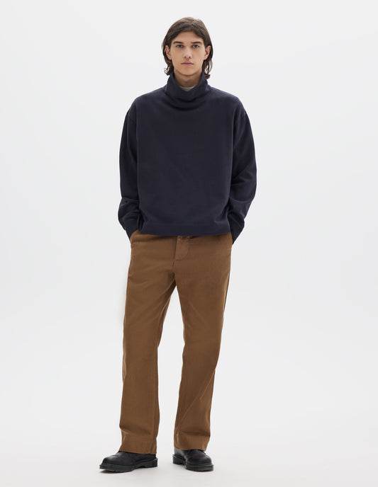 Full body image, front view of model wearing a dark navy loose funnel neck sweatshirt with single rib on cuffs and flatlock stitching on seams. Styled with brown cotton trousers and black leather shoes.