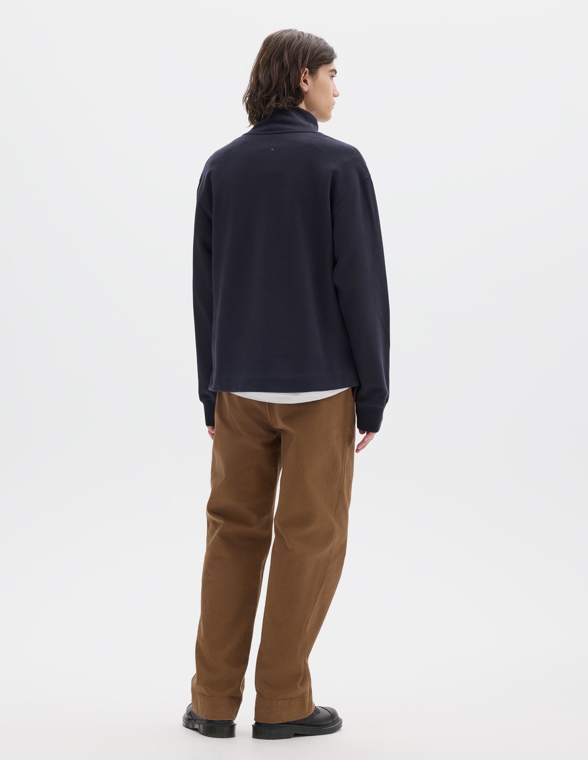 Full body image, front view of model wearing a dark navy loose funnel neck sweatshirt with single rib on cuffs and flatlock stitching on seams. Styled over a  white t shirt, with brown cotton trousers and black leather shoes.