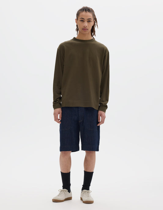 Full body image, front view of model wearing a brownish green cotton t shirt, with long sleeves, a deep ribbed collar and ribbed triangle insert on the shoulder. Styled with dark blue relaxed denim twill shorts, black socks and off white suede trainers.