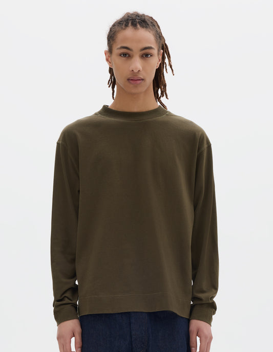 Cropped body image, front view of model wearing a brownish green cotton t shirt, with long sleeves, a deep ribbed collar and ribbed triangle insert on the shoulder. Styled with dark blue relaxed denim twill shorts.