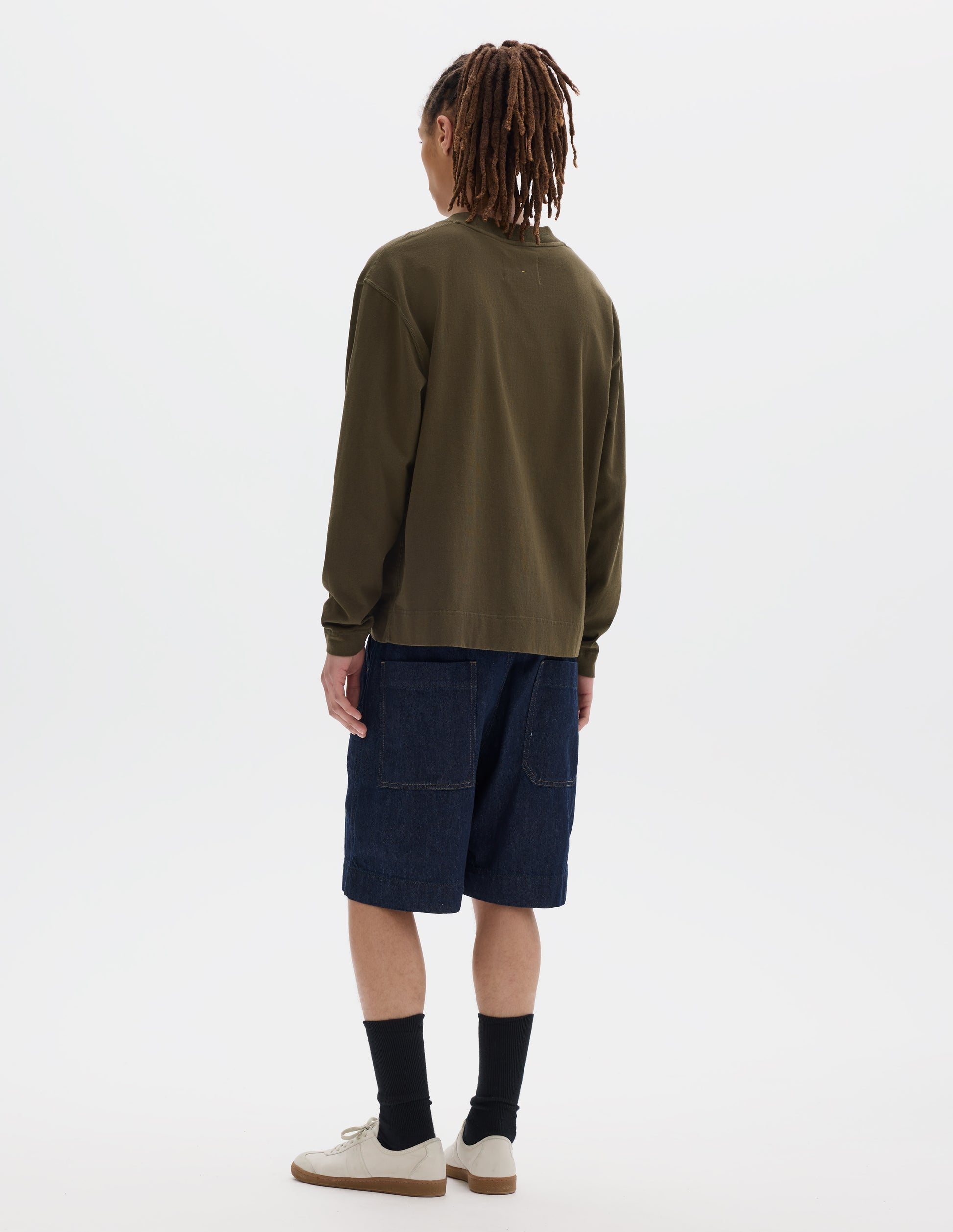 Full body image, back view of model wearing a brownish green cotton t shirt, with long sleeves, a deep ribbed collar and ribbed triangle insert on the shoulder. Styled with dark blue relaxed denim twill shorts, black socks and off white suede trainers.
