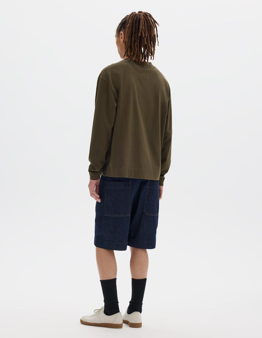 Full body image, back view of model wearing a brownish green cotton t shirt, with long sleeves, a deep ribbed collar and ribbed triangle insert on the shoulder. Styled with dark blue relaxed denim twill shorts, black socks and off white suede trainers.