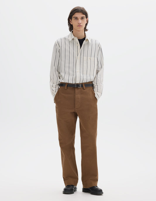 Full body image, front view of model wearing a wide stripe cotton shirt in off white and navy over a black t shirt, brown heavy cotton trousers with slant side pockets. Styled with a dark leather belt and black leather shoes.