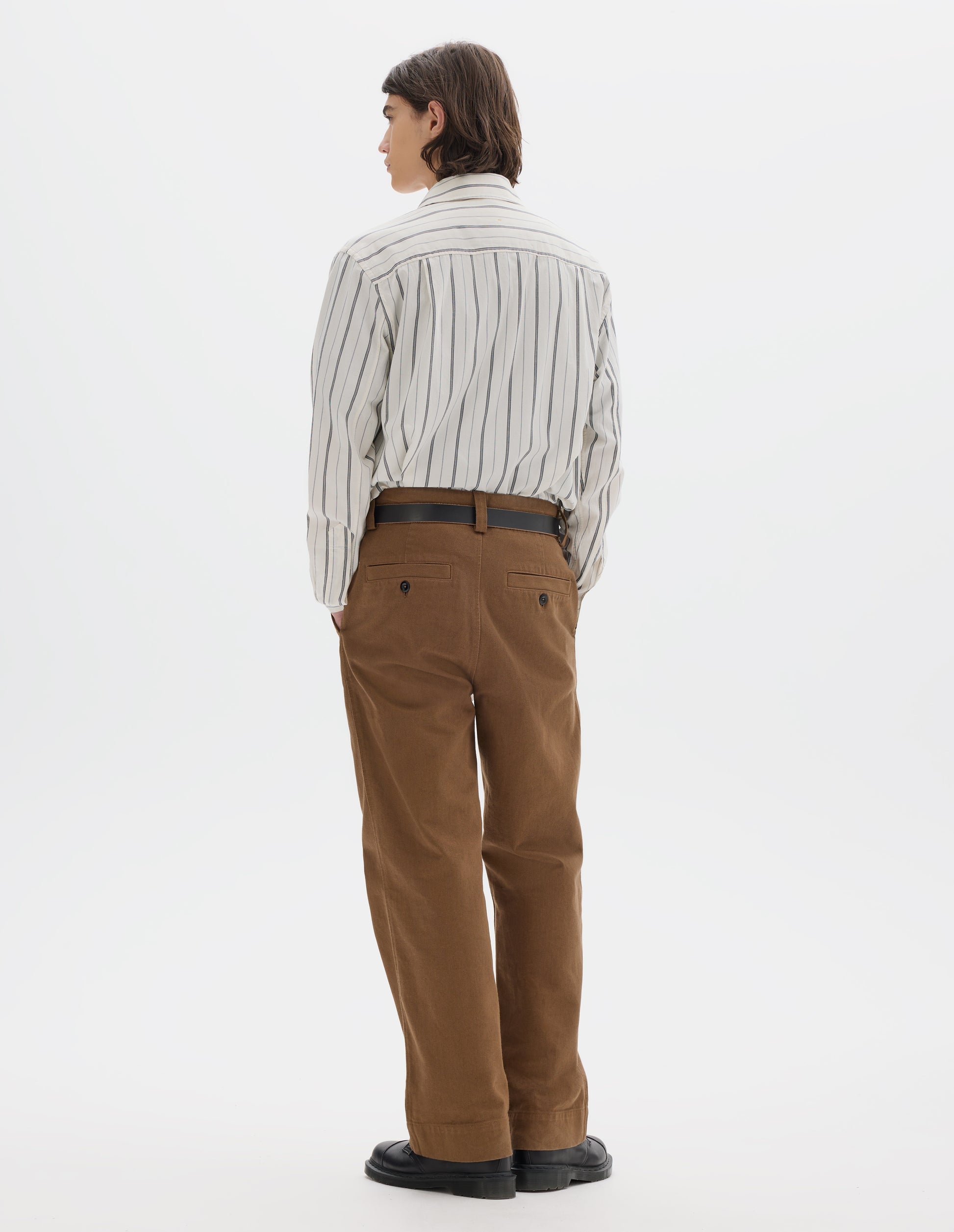 Full body image, back view of model wearing a wide stripe cotton shirt in off white and navy, brown heavy cotton trousers with slant side pockets and two back welt pockets with buttons. Styled with a dark leather belt and black leather shoes.