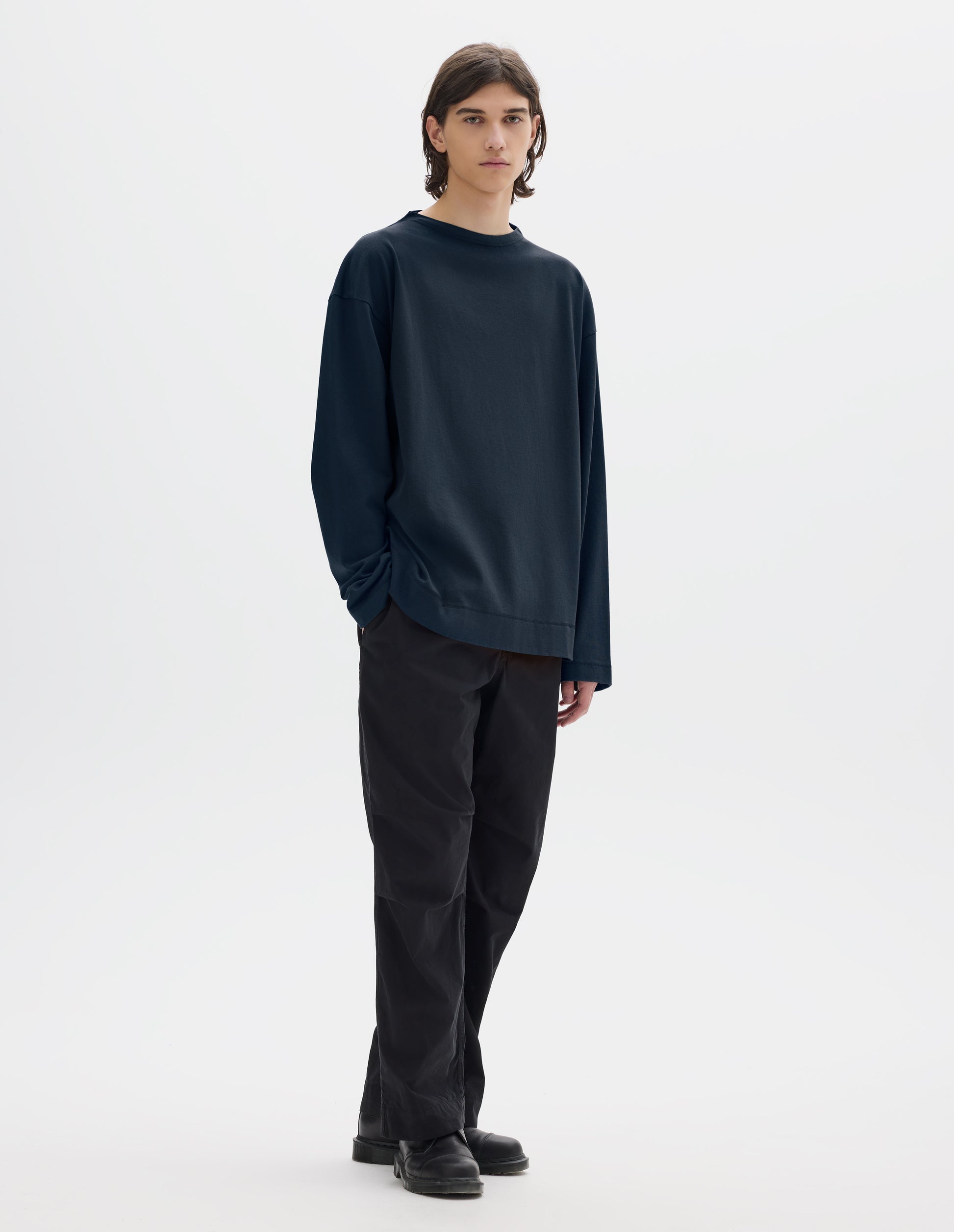 Full body image, front view of model wearing dark blue cotton t shirt, with long sleeves and flatlock stitching around cuffs and hem. Styled with black trousers and black shoes. 