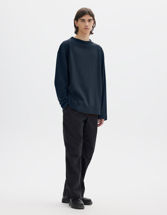Full body image, front view of model wearing dark blue cotton t shirt, with long sleeves and flatlock stitching around cuffs and hem. Styled with black trousers and black shoes. 