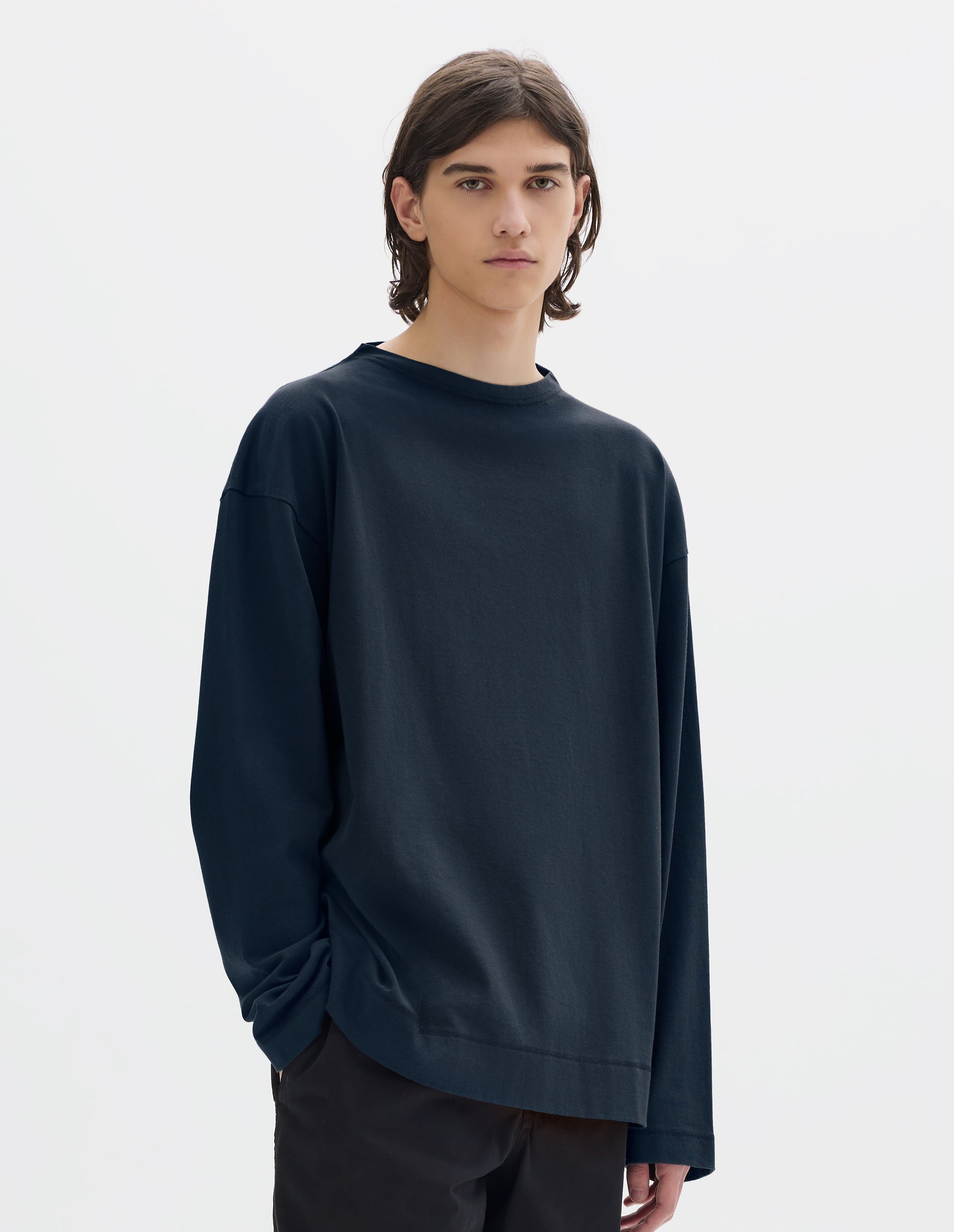 Cropped body image, front view of model wearing dark blue cotton t shirt, with long sleeves and flatlock stitching around cuffs and hem. Styled with black trousers.