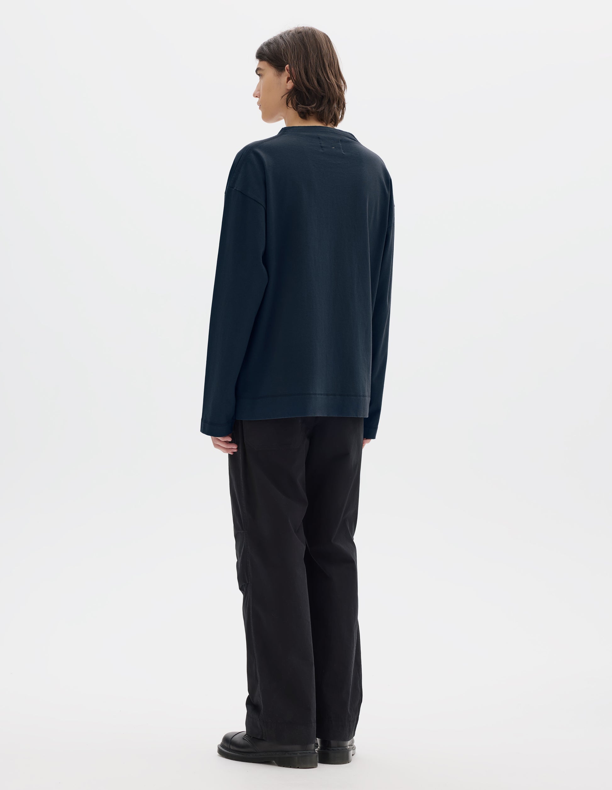 Full body image, back view of model wearing dark blue cotton t shirt, with long sleeves and flatlock stitching around cuffs and hem. Styled with black trousers and black shoes. 