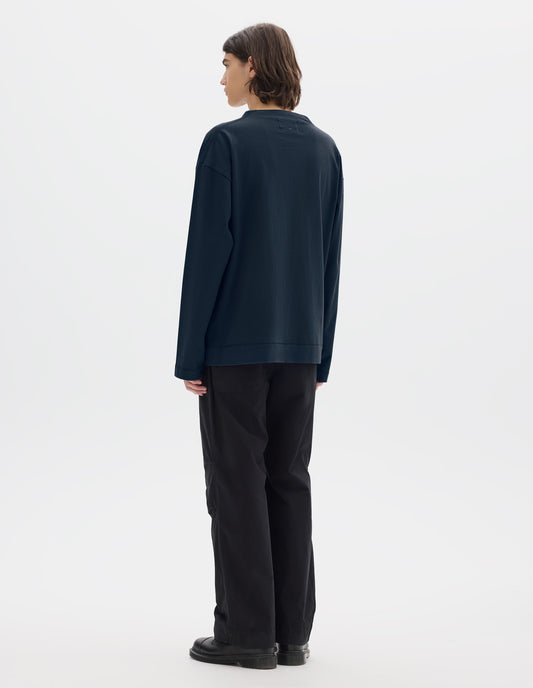 Full body image, back view of model wearing dark blue cotton t shirt, with long sleeves and flatlock stitching around cuffs and hem. Styled with black trousers and black shoes. 
