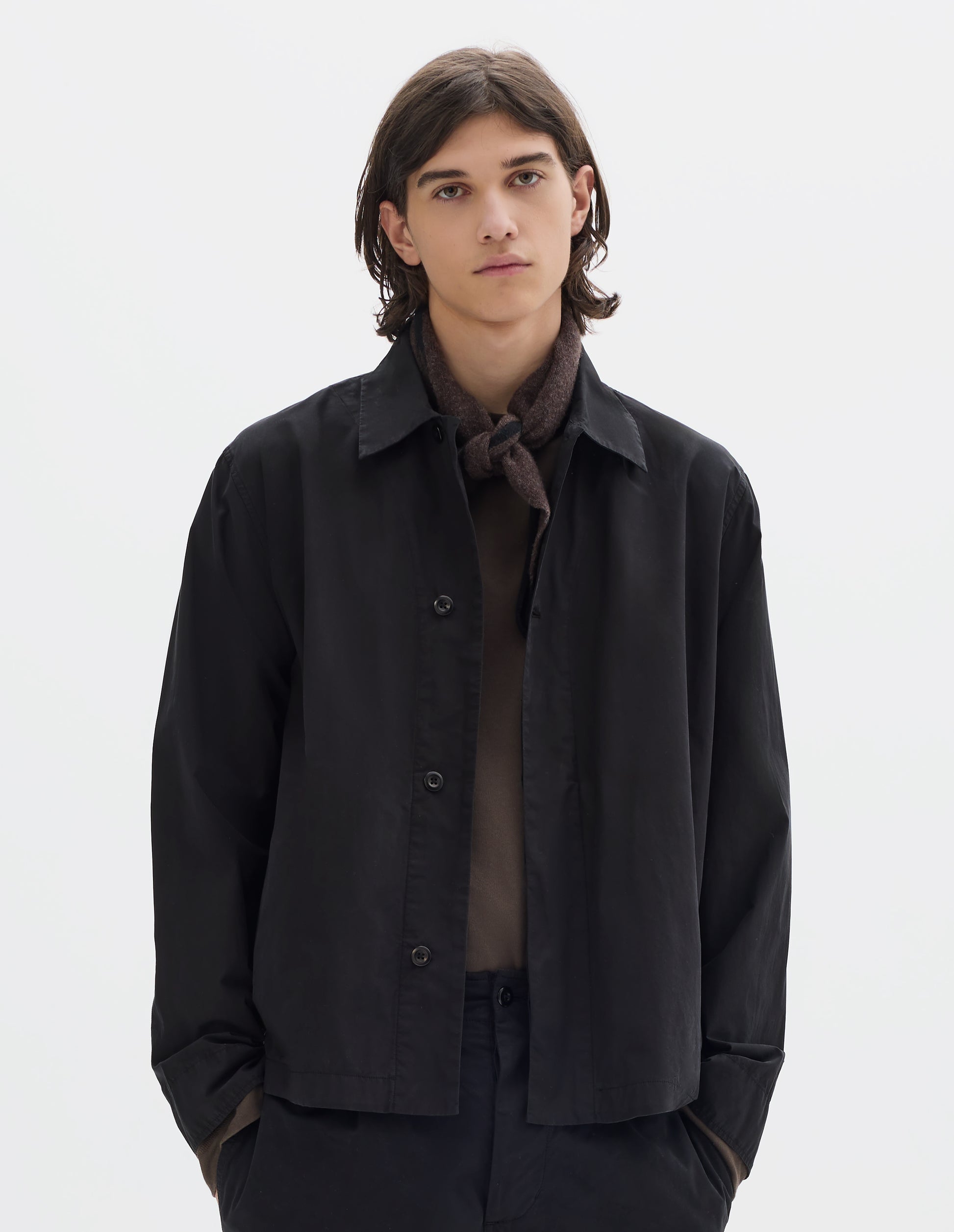 Cropped body image, front view of model wearing a black cotton overshirt, worn over a brown jersey. Styled with a brown wool triangle scarf with black edge and black cotton trousers.