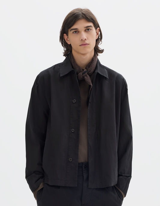 Cropped body image, front view of model wearing a black cotton overshirt, worn over a brown jersey. Styled with a brown wool triangle scarf with black edge and black cotton trousers.