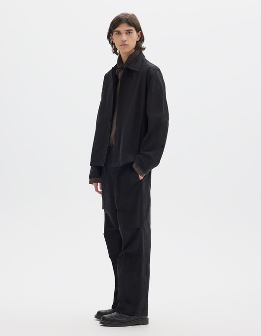 Full body image, front view of model wearing a relaxed boxy black overshirt with grown-on cuffs, and a stitch-through faced placket. Overshirt is worn open and styled over a brown jersey, brown black wool scarf and worn with relaxed cut black trousers and black leather shoes.