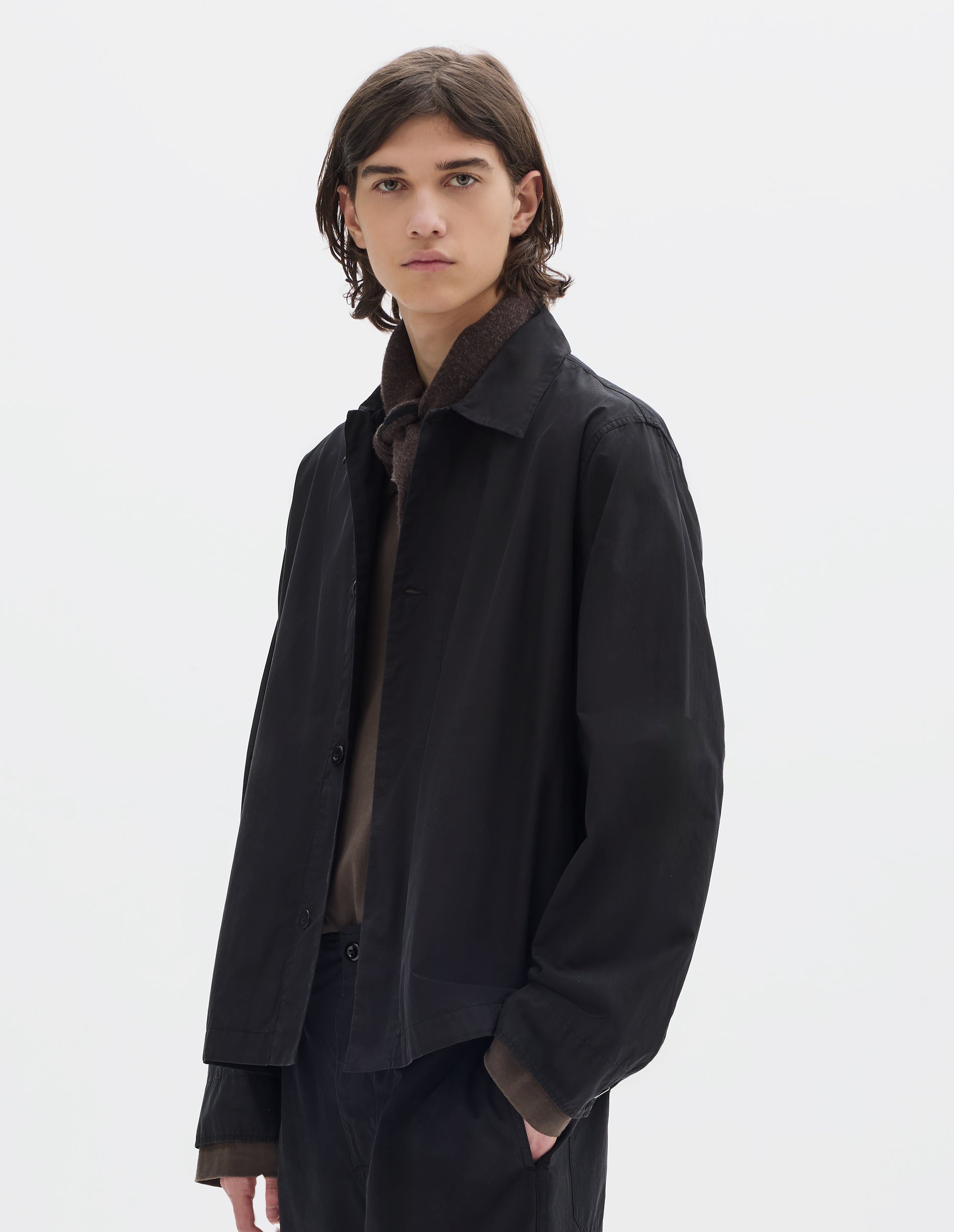 Cropped body image, front view of model wearing a relaxed boxy black overshirt with grown-on cuffs, and a stitch-through yoke. Overshirt is worn open and styled over a brown jersey, brown black wool scarf and worn with black trousers.