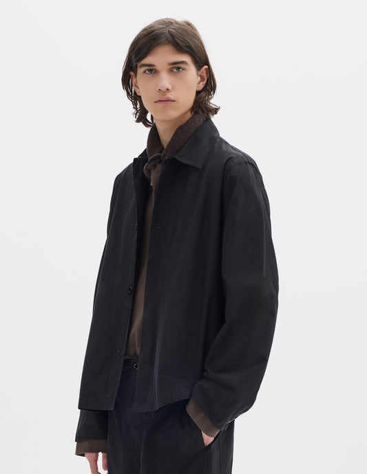 Cropped body image, front view of model wearing a relaxed boxy black overshirt with grown-on cuffs, and a stitch-through yoke. Overshirt is worn open and styled over a brown jersey, brown black wool scarf and worn with black trousers.