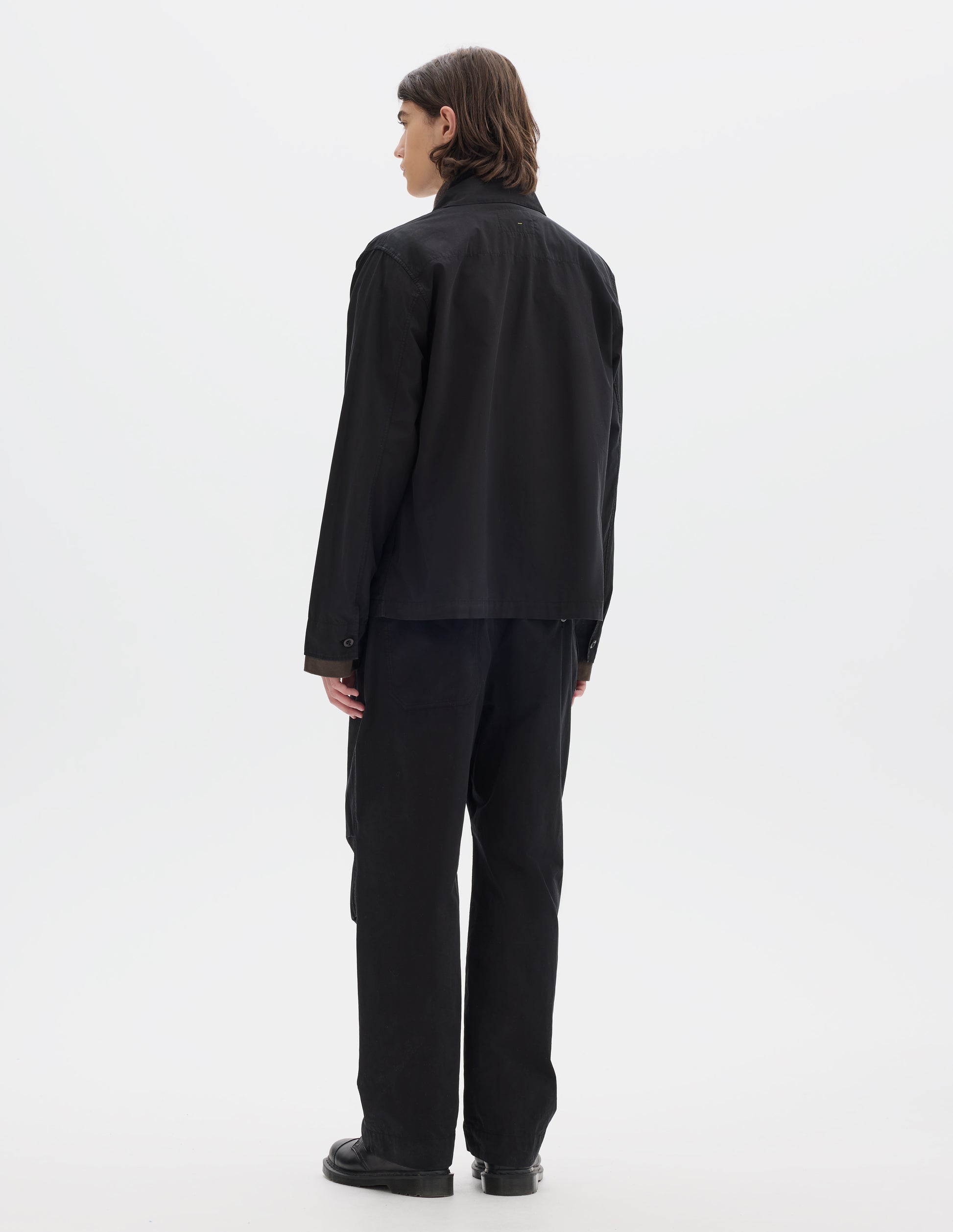 Full body image, back view of model wearing a relaxed boxy black overshirt with grown-on cuffs and a stitch-through yoke. Overshirt is worn over a brown long sleeve jersey, black trousers and black leather shoes.