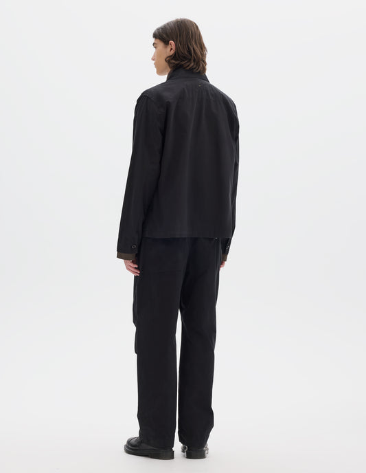 Full body image, back view of model wearing a relaxed boxy black overshirt with grown-on cuffs and a stitch-through yoke. Overshirt is worn over a brown long sleeve jersey, black trousers and black leather shoes.
