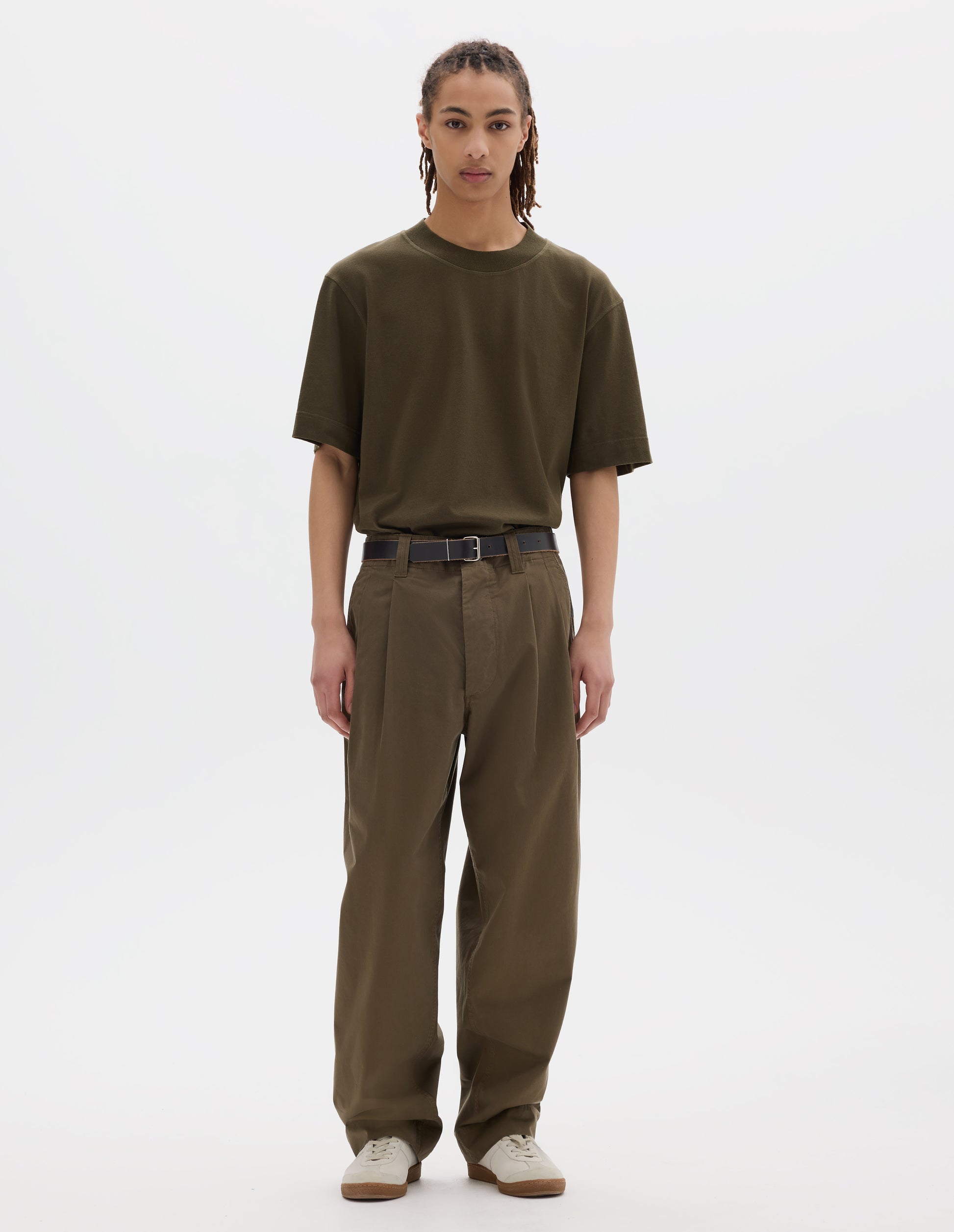 Full body image, front view of model wearing a green brown flatlock crew short sleeve T shirt which features a rib neck and flatlock stitch detailing on the seams. Styled with a dark leather belt, green brown pleated trousers with slant pockets and off white suede trainers.