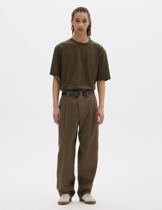 Full body image, front view of model wearing a green brown flatlock crew short sleeve T shirt which features a rib neck and flatlock stitch detailing on the seams. Styled with a dark leather belt, green brown pleated trousers with slant pockets and off white suede trainers.
