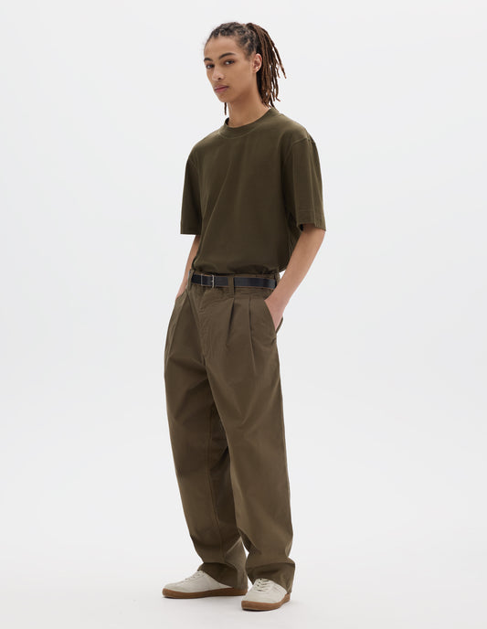 Full body image, front view of model wearing a green brown flatlock crew short sleeve T shirt which features a rib neck and flatlock stitch detailing on the seams. Styled with a dark leather belt, green brown pleated trousers with slant pockets and off white suede trainers.