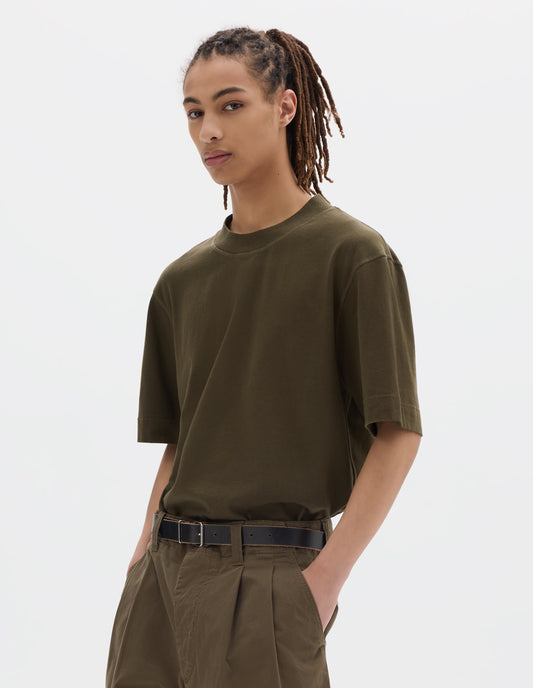 Cropped body image, front view of model wearing a green brown flatlock crew short sleeve T shirt which features a rib neck and flatlock stitch detailing on the seams. Styled with a dark leather belt and green brown pleated trousers.