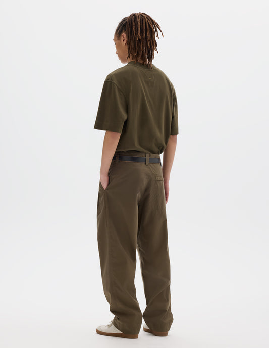 Full body image, back view of model wearing agreen brown flatlock crew short sleeve T shirt which features a rib neck and flatlock stitch detailing on the seams. Styled with a dark leather belt and green brown trousers with stitch-through back pocket with concealed zip fastening and off white suede trainers.