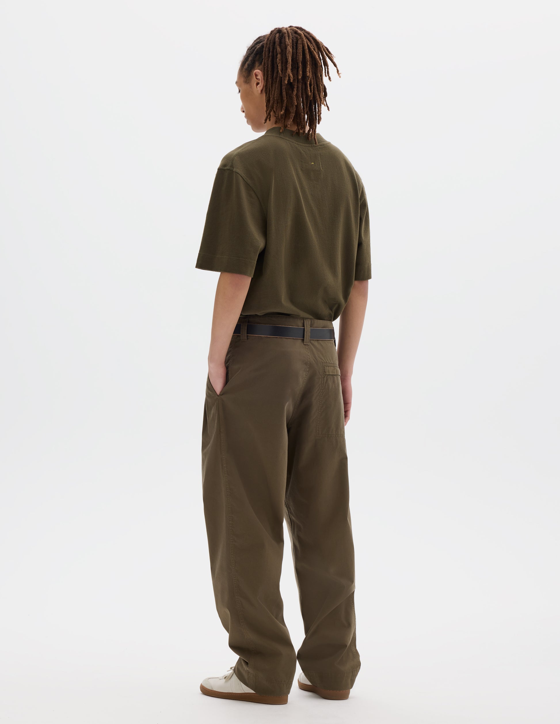 Full body image, back view of model wearing agreen brown flatlock crew short sleeve T shirt which features a rib neck and flatlock stitch detailing on the seams. Styled with a dark leather belt and green brown trousers with stitch-through back pocket with concealed zip fastening and off white suede trainers.