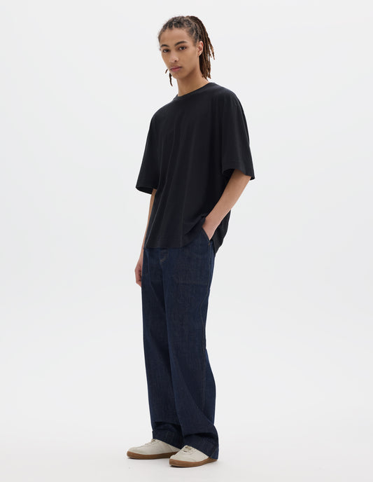 Full body image, front view of model wearing boxy black t-shirt with herringbone tape detailing on inside neck and vent split hem. Styled with dark indigo denim trousers and white suede trainers.
