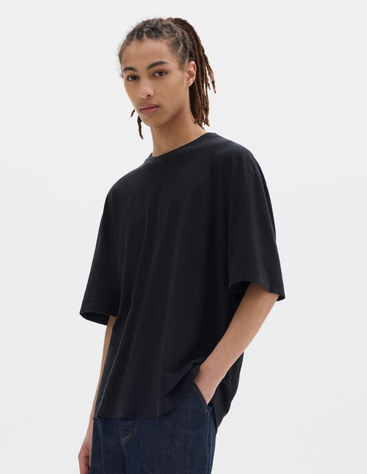 Cropped body image, front view of model wearing boxy black t-shirt with herringbone tape detailing on inside neck and vent split hem. Styled with dark indigo denim trousers.
