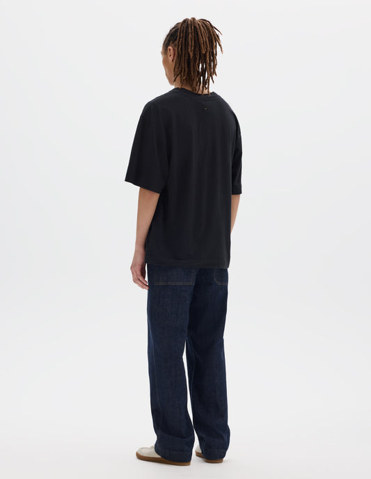 Full body image, back view of model wearing boxy black t-shirt with herringbone tape detailing on inside neck and vent split hem. Styled with dark indigo denim trousers and white suede trainers.