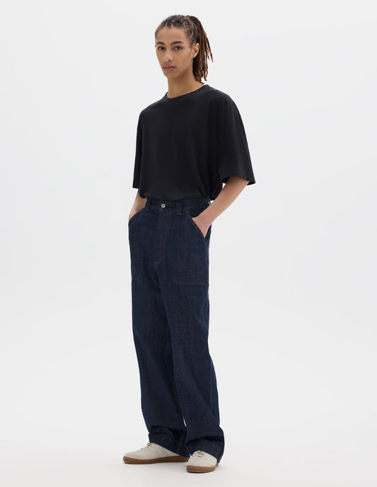 Full body image, front view of model wearing an indigo blue denim twill jeans with metal button tab waist adjusters, metal button fly, belt loops and slant stitch-through pockets. Styled with black cotton linen short sleeve T shirt and white suede trainers.