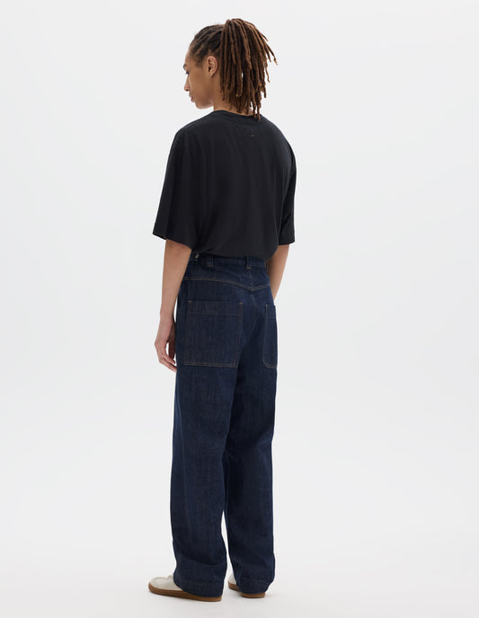 Full body image, back view of model wearing an indigo blue denim twill jeans with metal button tab waist adjusters, belt loops and patch and hammer pocket on back of trousers. Styled with black cotton linen short sleeve T shirt and white suede trainers.