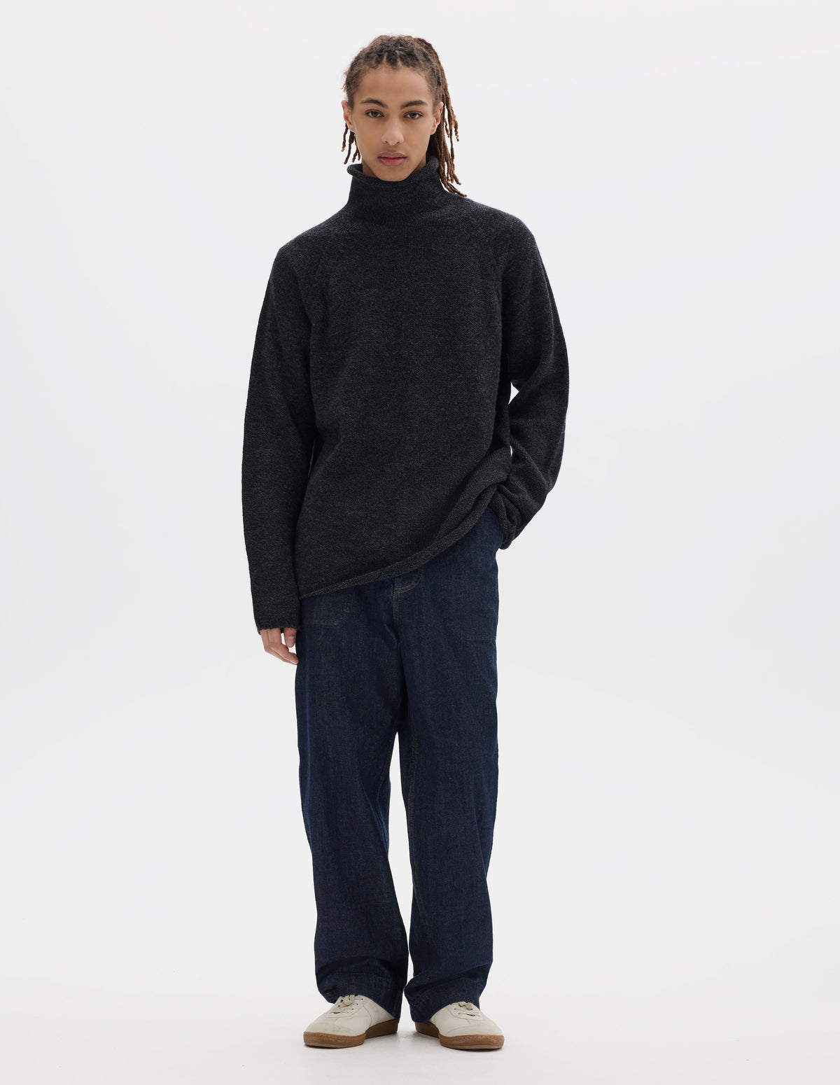 Full body image, front view of model wearing an off black high neck wool sweater with raglan sleeves, rolled cuffs and hem. Styled with dark blue denim trousers and off white suede trainers.