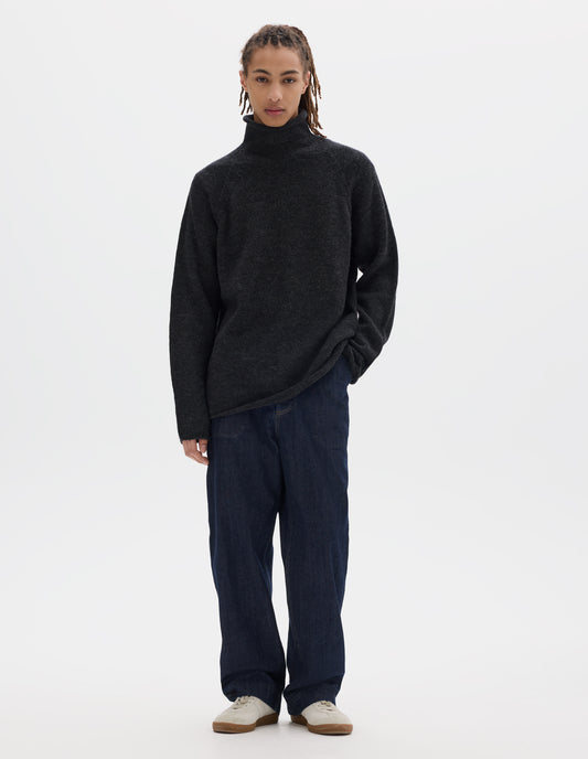 Full body image, front view of model wearing an off black high neck wool sweater with raglan sleeves, rolled cuffs and hem. Styled with dark blue denim trousers and off white suede trainers.