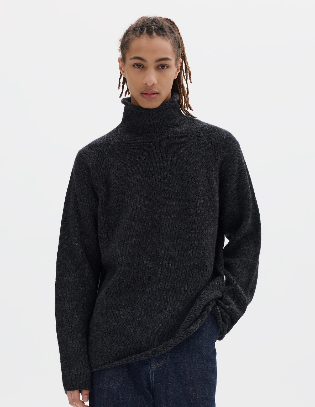 Cropped body image, front view of model wearing an off black high neck wool sweater with raglan sleeves, rolled cuffs and hem. Styled with dark blue denim trousers.
