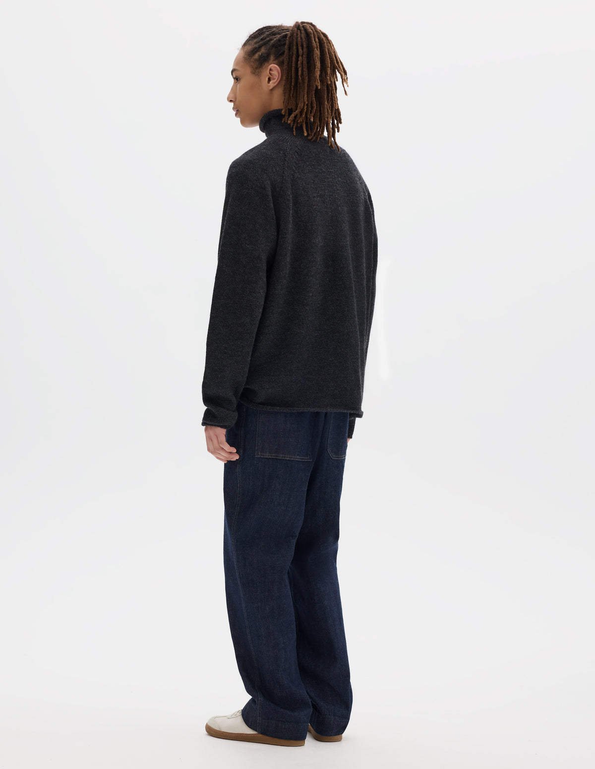 Full body image, back view of model wearing an off black high neck wool sweater with raglan sleeves, rolled cuffs and hem. Styled with dark blue denim trousers and off white suede trainers.