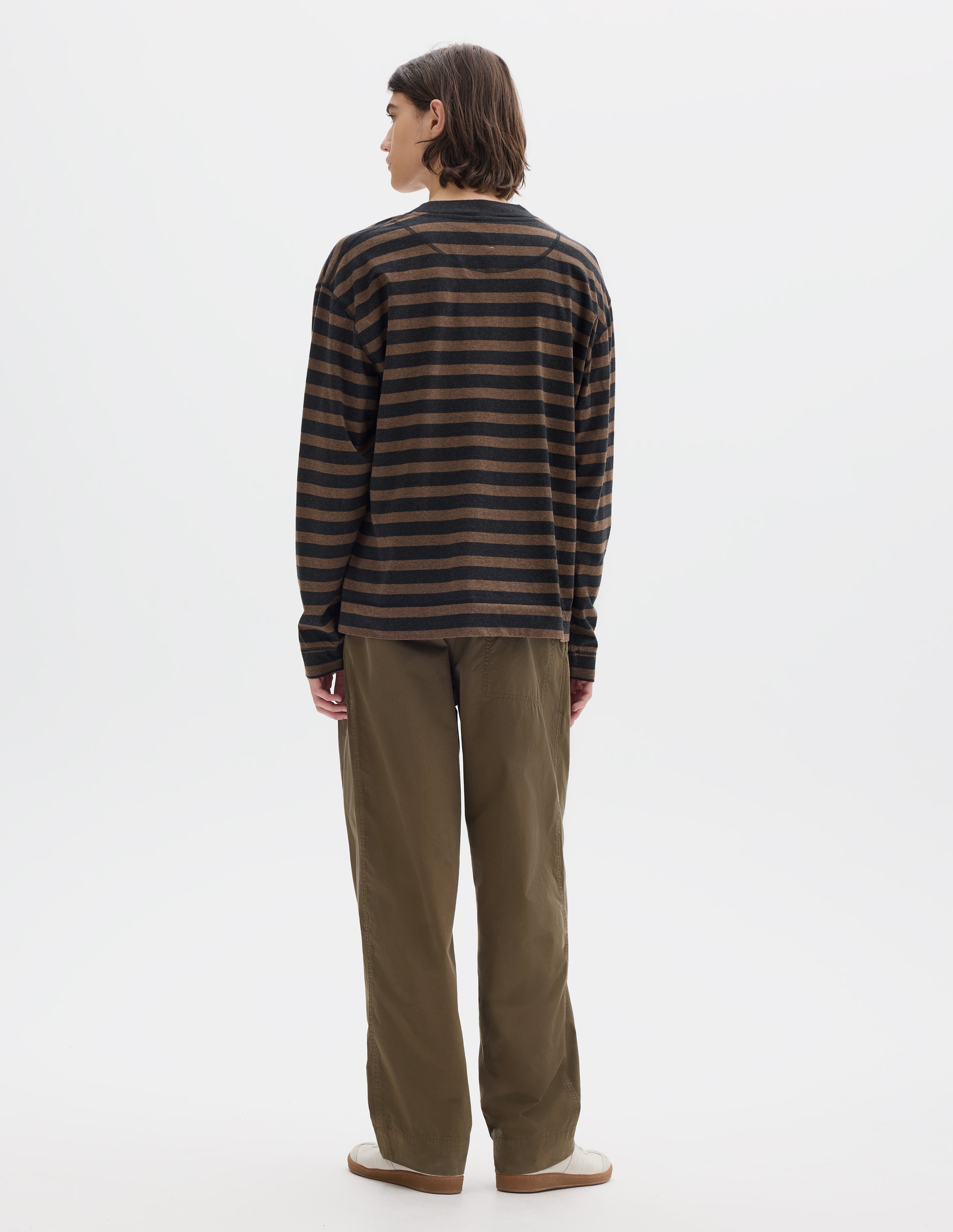 Full body image, back view of model wearing black and brown stripe crew neck long sleeve jersey, rib trim on cuff and neck, flatlock stitch seam detailing. Styled with khaki trousers and white shoes.