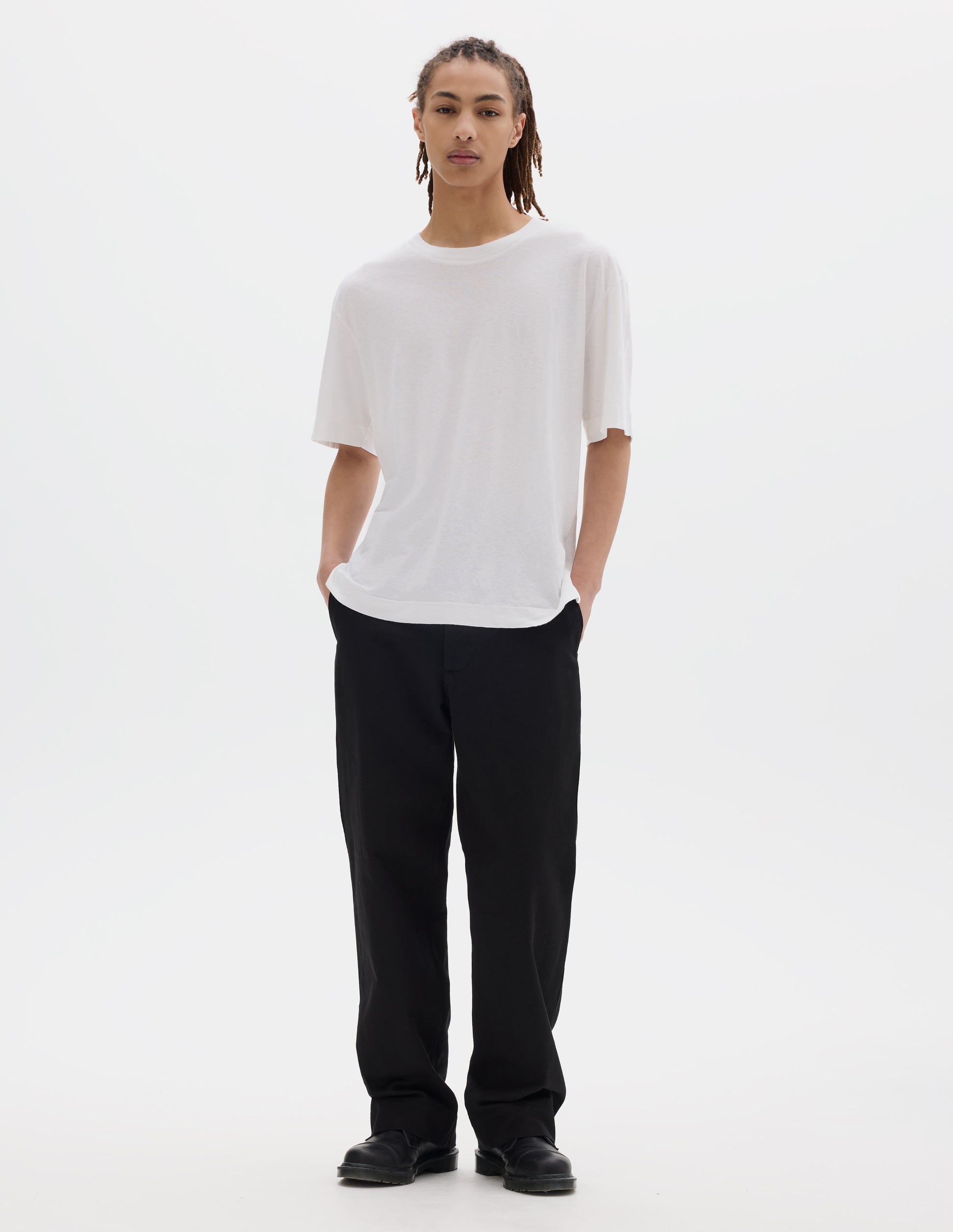 Full body image, front view of model wearing a boxy white t-shirt with herringbone tape detailing on inside neck and vent split hem. Styled with black cotton trousers and black leather shoes.
