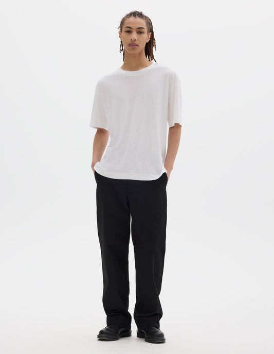 Full body image, front view of model wearing a boxy white t-shirt with herringbone tape detailing on inside neck and vent split hem. Styled with black cotton trousers and black leather shoes.