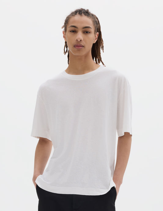 Cropped body image, front view of model wearing a boxy white t-shirt with herringbone tape detailing on inside neck and vent split hem. Styled with off black cotton trousers.