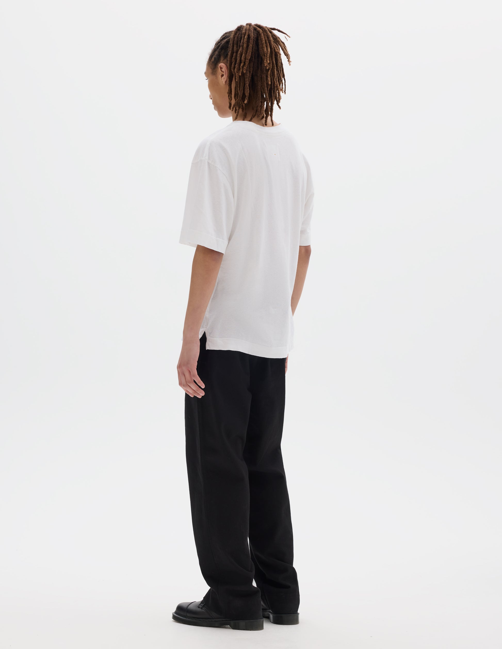 Full body image, back view of model wearing a boxy white t-shirt with vent split hem. Styled with black cotton trousers and black leather shoes.