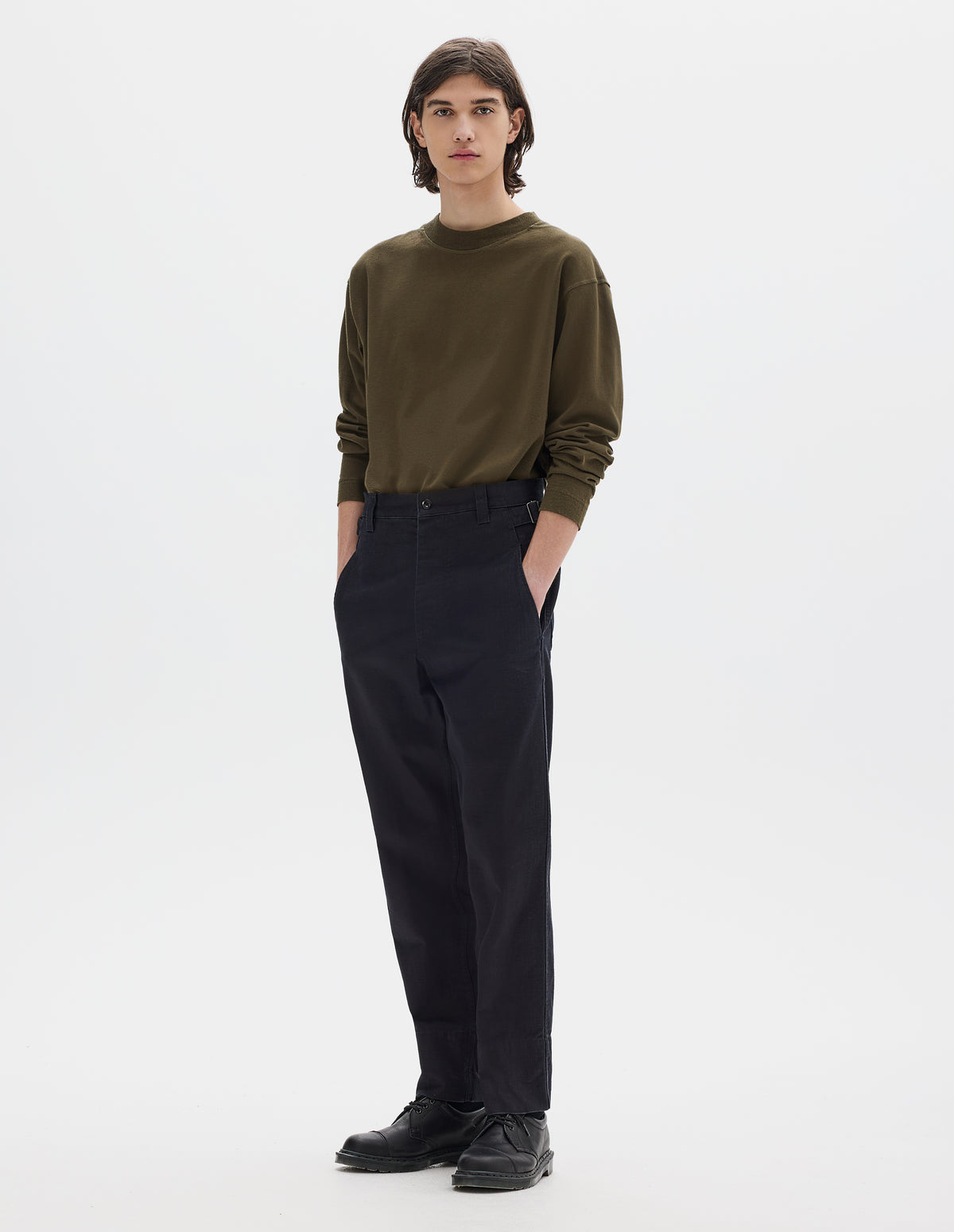 Full body image, front view of model wearing dark blue trousers with belt loops and side cinches, slant pockets, zip fly and corozo button fastening, deep stitch hem. Styled with brown long sleeve t-shirt tucked in and black leather shoes.